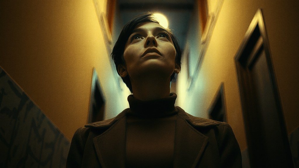 The image shows a person standing in a dimly lit hallway, looking upward with a contemplative expression.