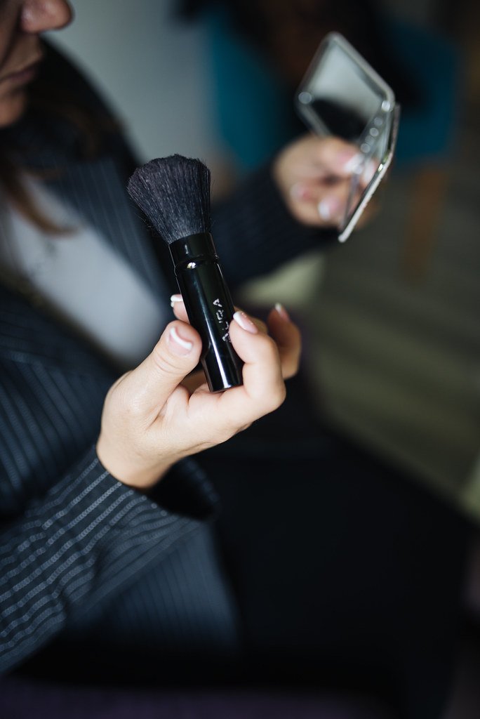 A person holds a black makeup brush in one hand and a small mirro