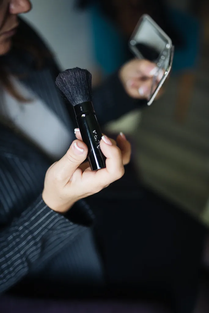 A person holds a black makeup brush in one hand and a small mirro