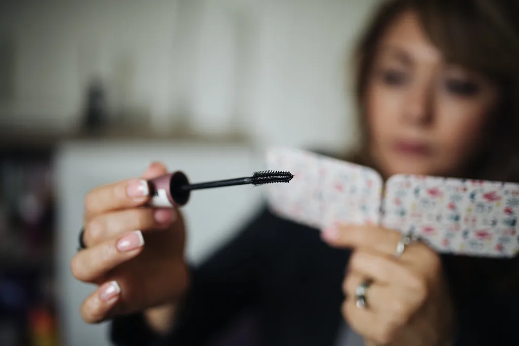 A woman carefully applies mascara to her lashes while holding a floral-patterned mirror in a cozy indoor setting