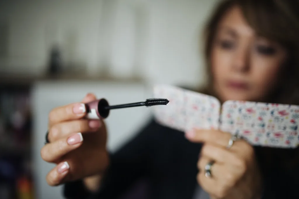 A woman carefully applies mascara to her lashes while holding a floral-patterned mirror in a cozy indoor setting