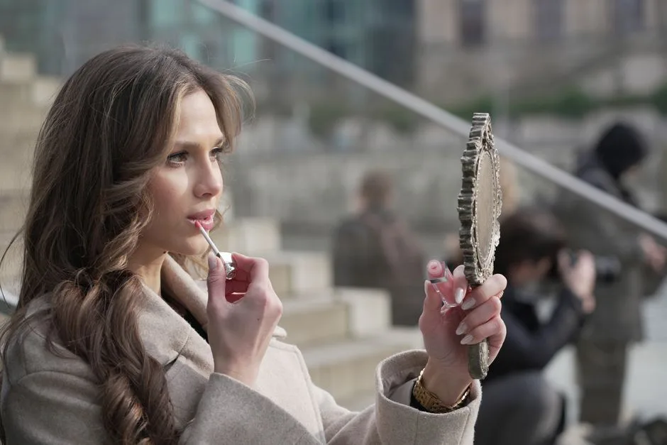 A fashionable woman applies lipstick while gazing into a hand mirror on a Berlin street.
