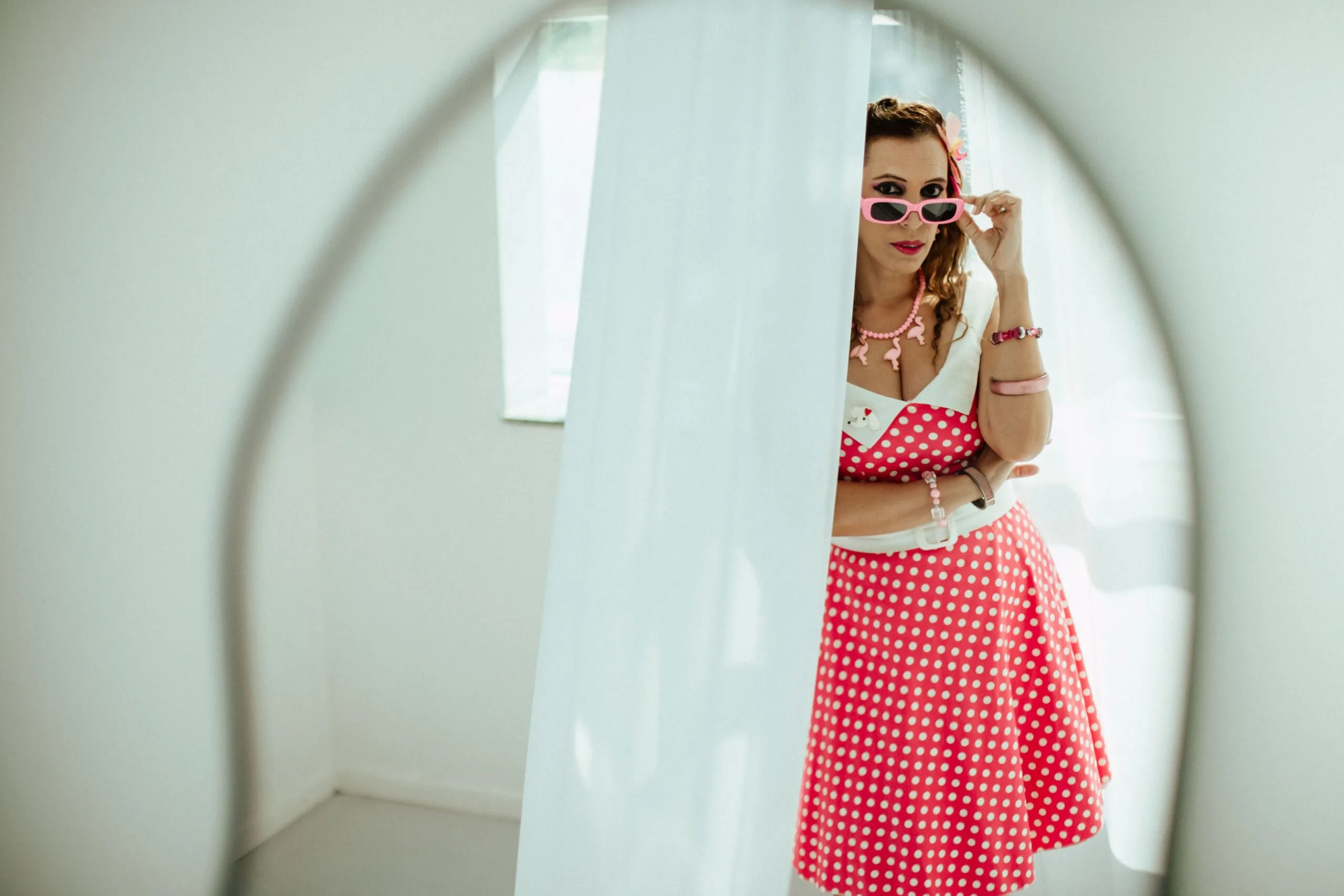 Stylish woman in pink and white polka dot dress posing with sunglasses indoors.
