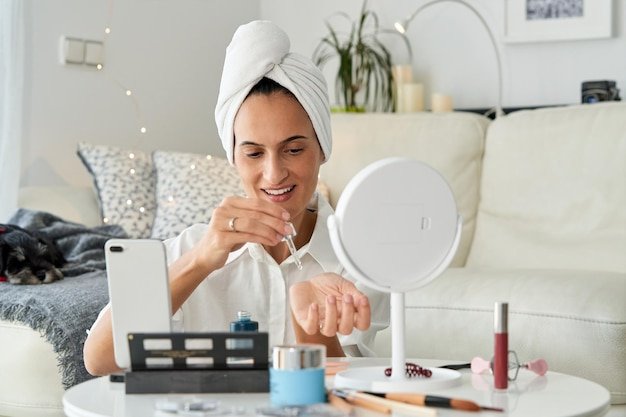 Cheerful female vlogger with turban of towel on head applying cosmetic moisturizing serum while recording skin care tutorial video on smartphone for vlog in social media