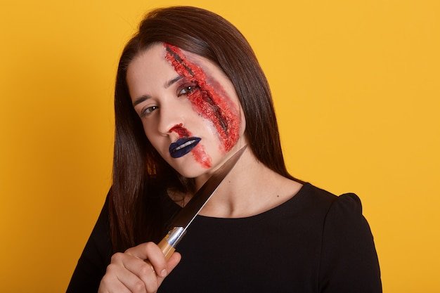 Indoor shot of brunette female with knife near her neck and bloody wound on her face