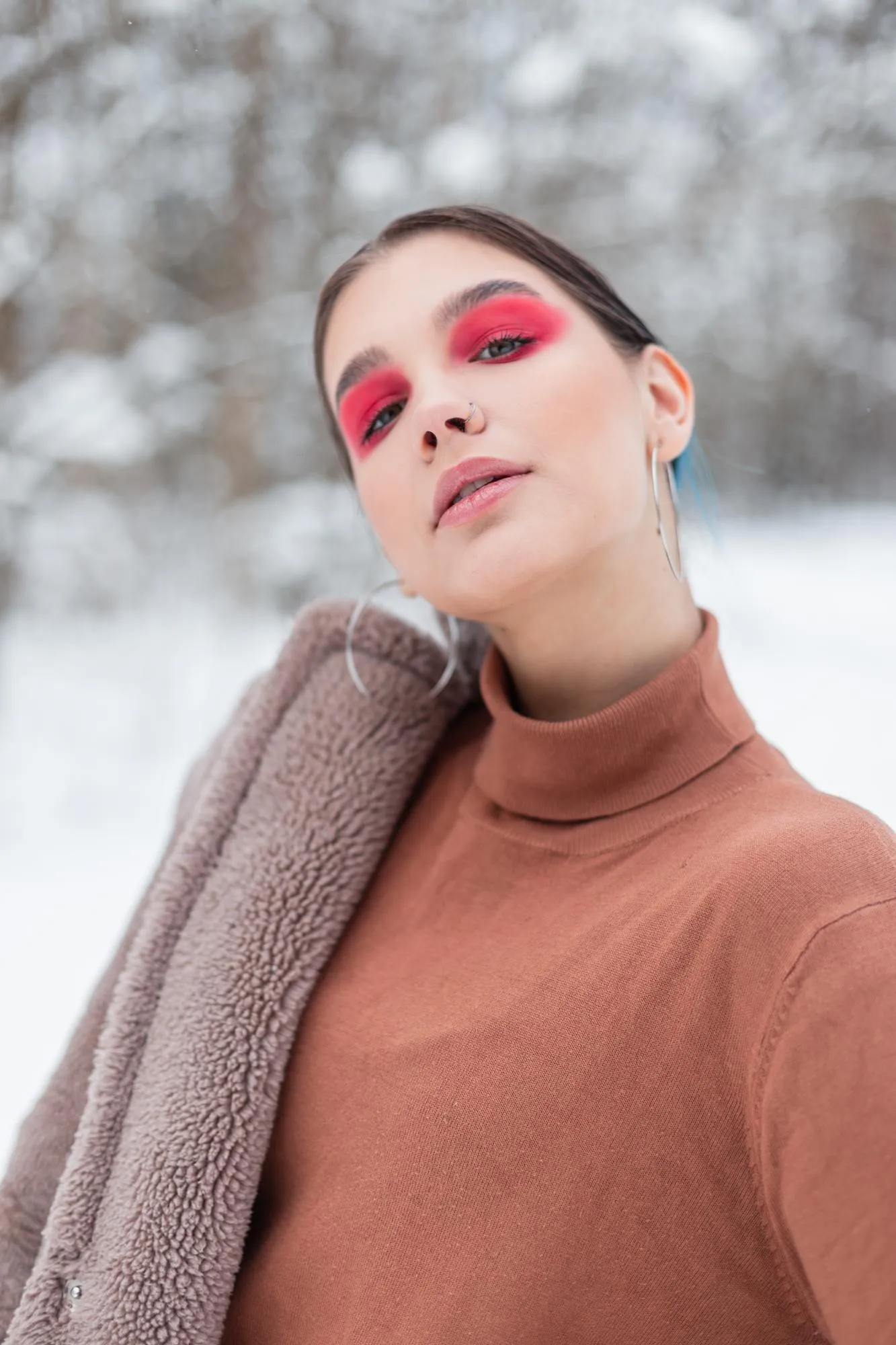 Vogue portrait of a beautiful model girl with bright colored pink makeup and eye shadows in fashionable clothes with a sweater, coat and scarf in a snowy forest outdoors