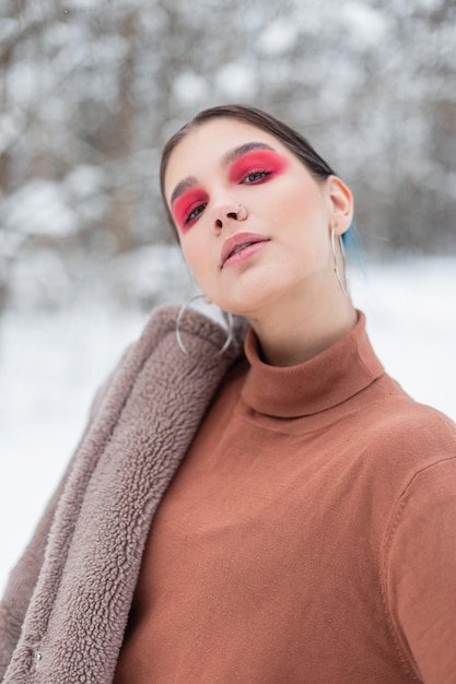 Vogue portrait of a beautiful model girl with bright colored pink makeup and eye shadows in fashionable clothes with a sweater, coat and scarf in a snowy forest outdoors