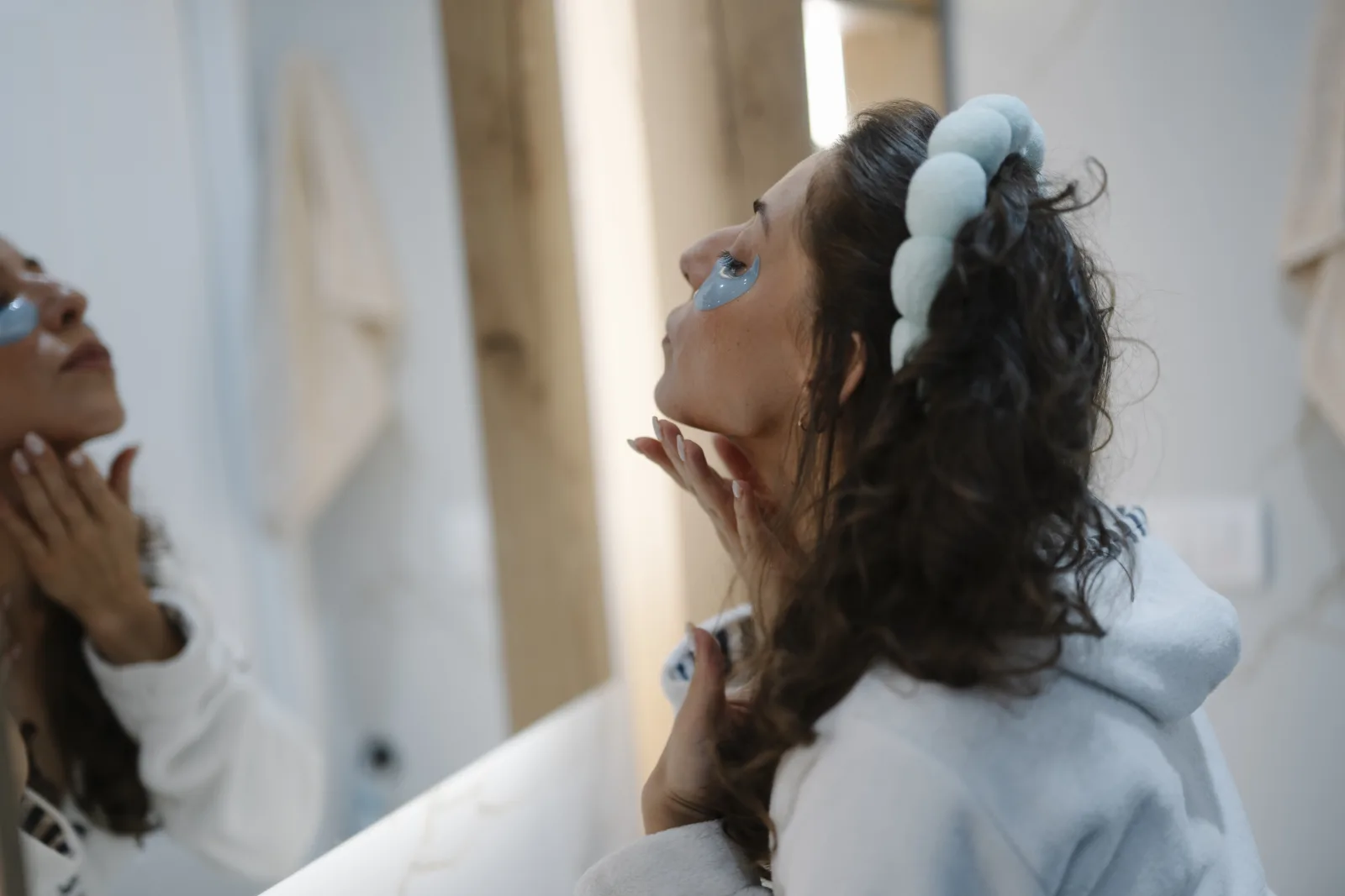 A woman is standing in front of a mirror in a bathroom. She is using a skincare product on her face while dressed in comfortable clothes. The setting shows a clean space with natural light.