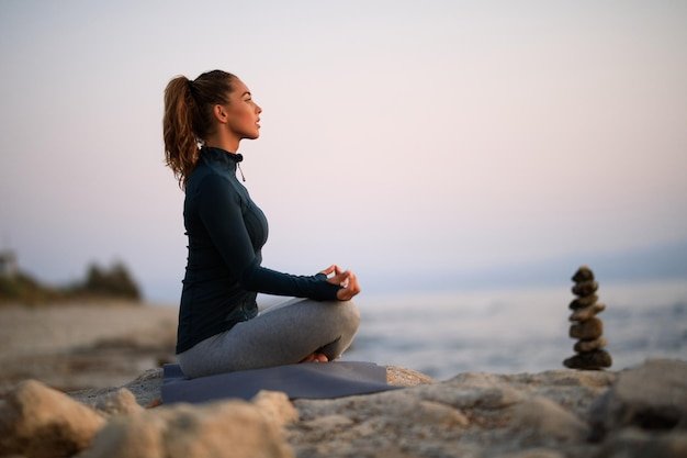 Athletic woman practicing Yoga and doing breathing exercise in lotus positing on a  beach rock.