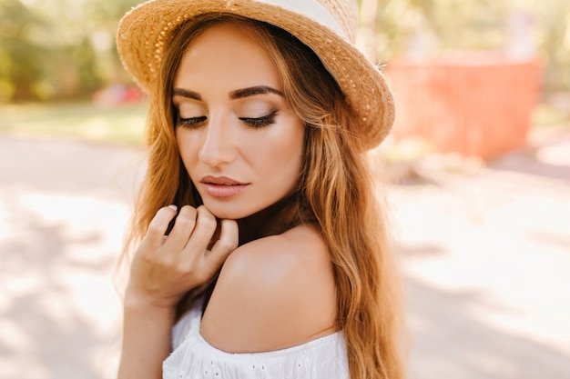 Close-up portrait of pensive woman with lightly-tanned skin and trendy nude make-up posing with eyes closed. Outdoor photo of lady in vintage hat touching chin and looking down.