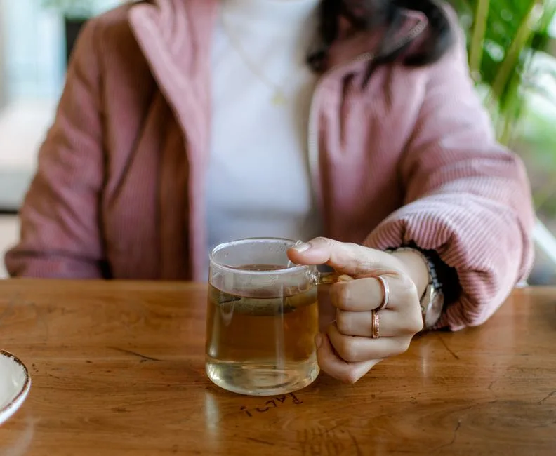 A woman holds a clear glass of herbal tea at a wooden table in a cozy cafe setting.