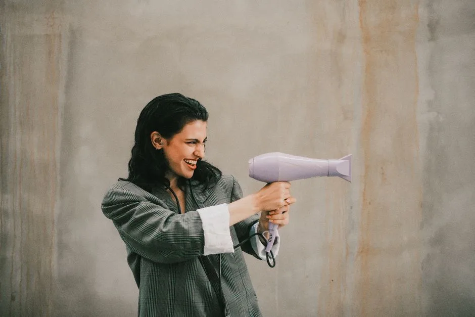 Playful woman in casual attire posing with a hairdryer against an industrial backdrop.