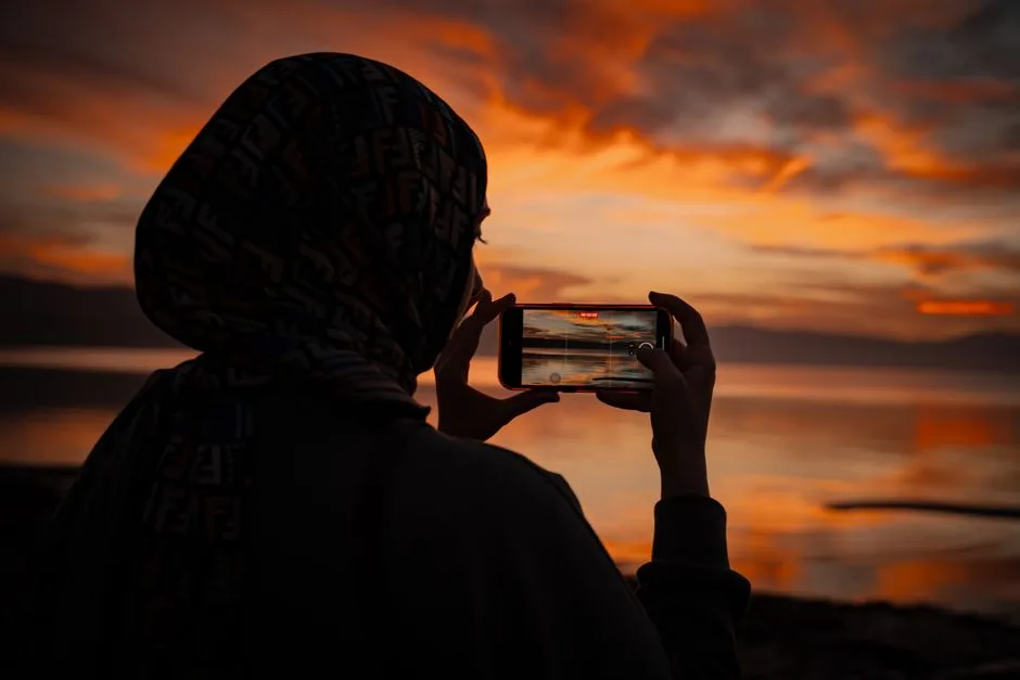Silhouette of a person capturing a vibrant sunset over a serene lake with a smartphone