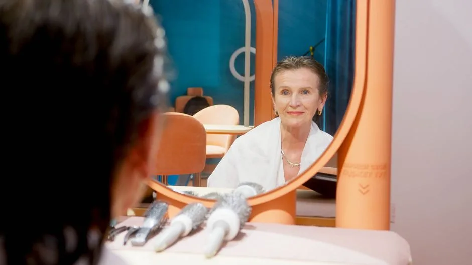 Senior woman looking at her reflection in a salon mirror, smiling with hairbrushes on counter.