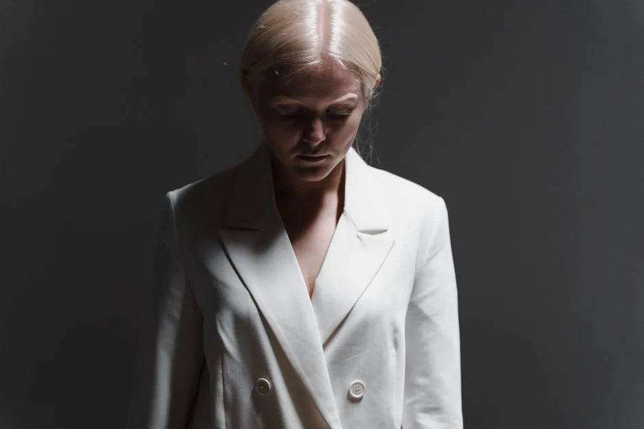 Studio portrait of a woman with blonde hair looking down, wearing a white coat.