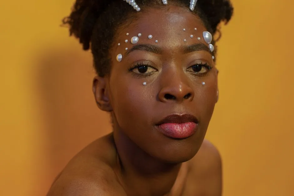 Close-up portrait of a woman with pearl accents on her face against a yellow background.