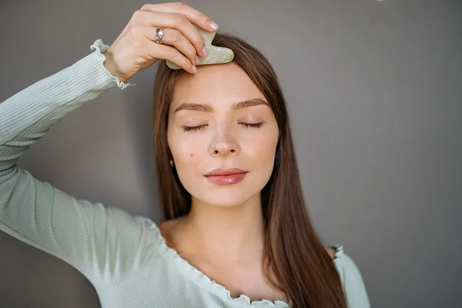 A woman relaxes with a jade gua sha tool for a facial skincare routine against a gray background.
