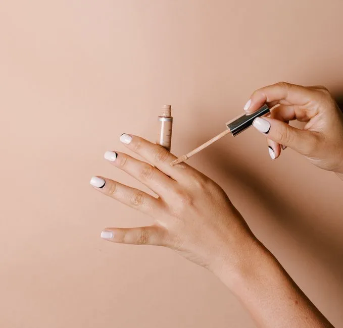 Elegantly manicured hands applying liquid concealer on skin against a neutral background.