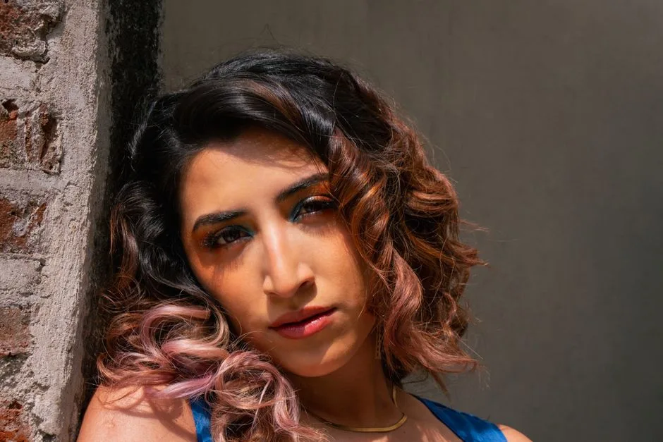 Close-up portrait of a woman with curly hair, highlighted by natural sunlight against a rustic brick wall.