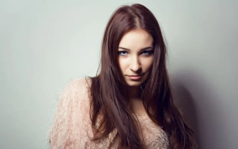 Close-up portrait of a young woman with long brown hair and a stylish outfit against a neutral background.
