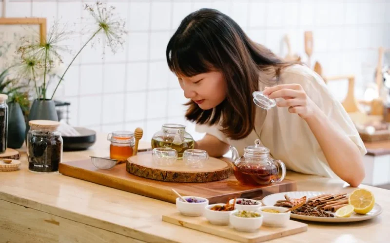 Woman preparing herbal tea with natural ingredients in a kitchen setting, focusing on aroma and presentation.