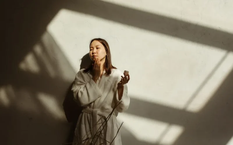 An Asian woman in a robe applies facial cream in soft, natural sunlight indoors.