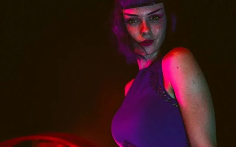 A fashionable woman with purple hair poses confidently in a colorful, low-light setting.
