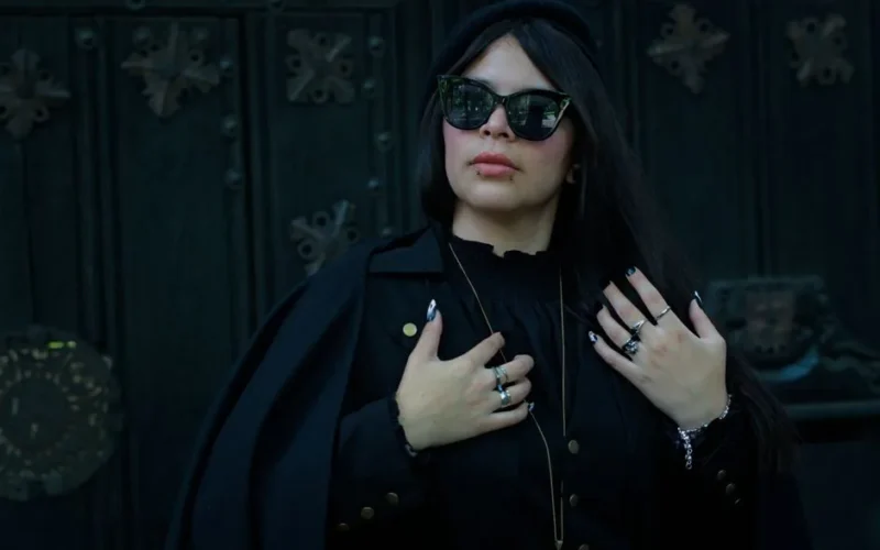 A fashionable woman in gothic attire poses confidently against a dark ornate background.
