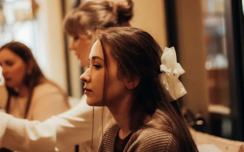 Profile of a young woman with a white bow in her hair, in a cozy indoor environment.