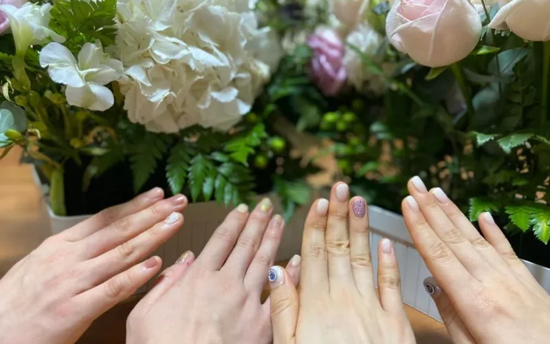 Group of hands with varied nail art surrounded by lush flower arrangements in pastel tones.