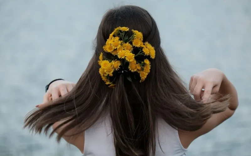 A woman with yellow flowers in her brunette hair, standing by a serene water body.