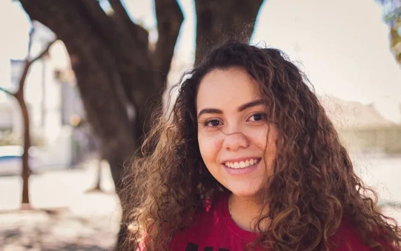 Portrait of a smiling young woman with curly hair in a sunny outdoor setting.