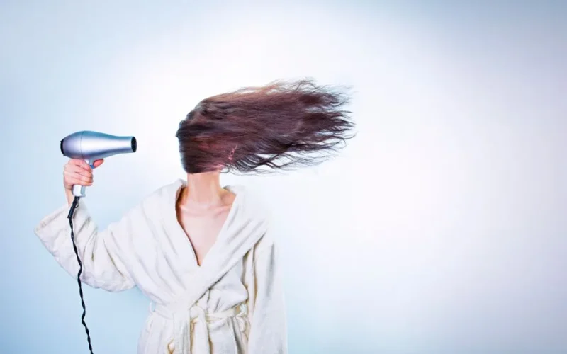 Woman in a bathrobe using a hairdryer, hair flying wildly. Indoor setting.
