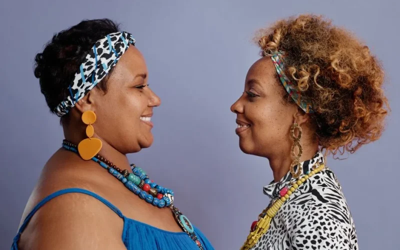 Side view of two women with vibrant accessories and headbands facing each other against a purple background.