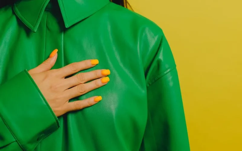 Close-up of a woman's hand with yellow nails on a green leather jacket against a yellow background.