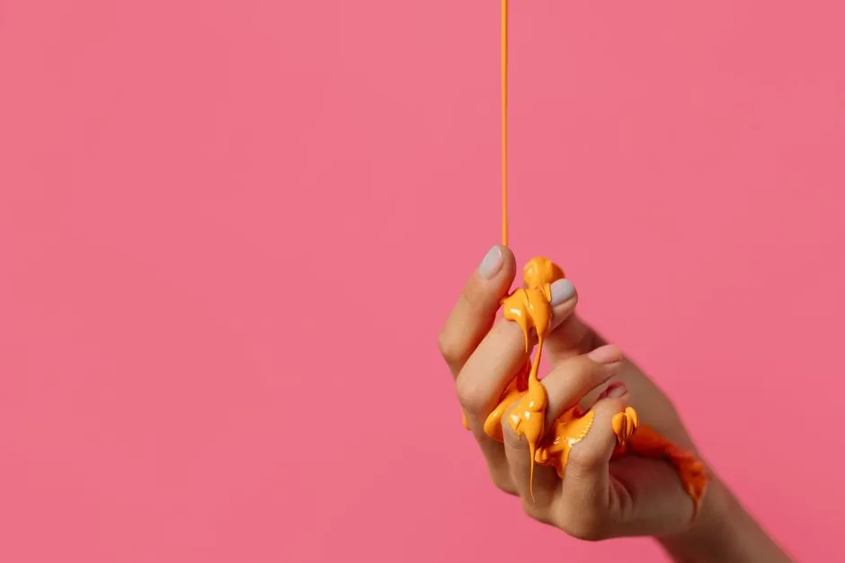 A close-up of a hand with dripping orange paint on a pink background.