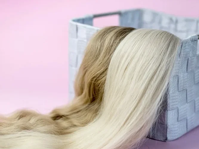 Close-up of blond and brown hair extensions draped over a woven basket, on a pink background.