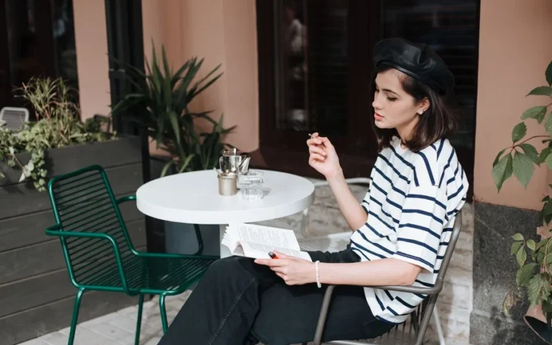 Stylish woman in stripes relaxes with a book and cigarette at an outdoor cafe.