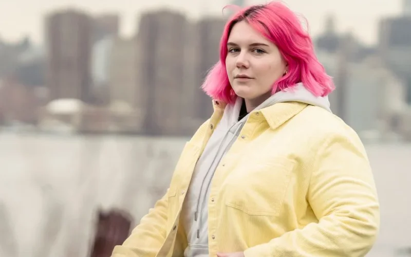 A woman with pink hair stands outdoors wearing a yellow jacket, with a city skyline in the background.