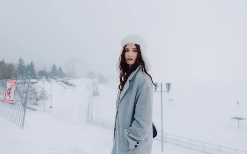 A fashionable woman wearing a grey coat and white hat standing in a snowy urban setting.