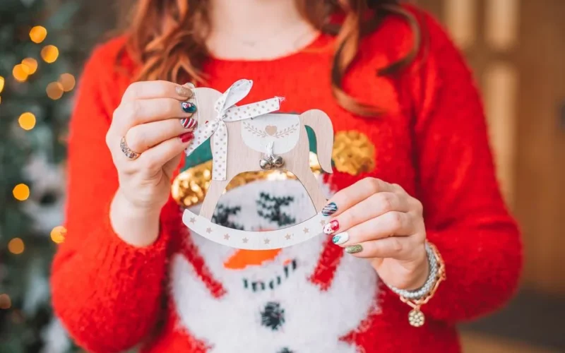 Close-up of a woman in a festive sweater holding a Christmas ornament, showcasing her nail art.