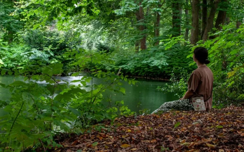 A woman sits on a riverbank surrounded by vibrant green trees, reflecting peacefully.