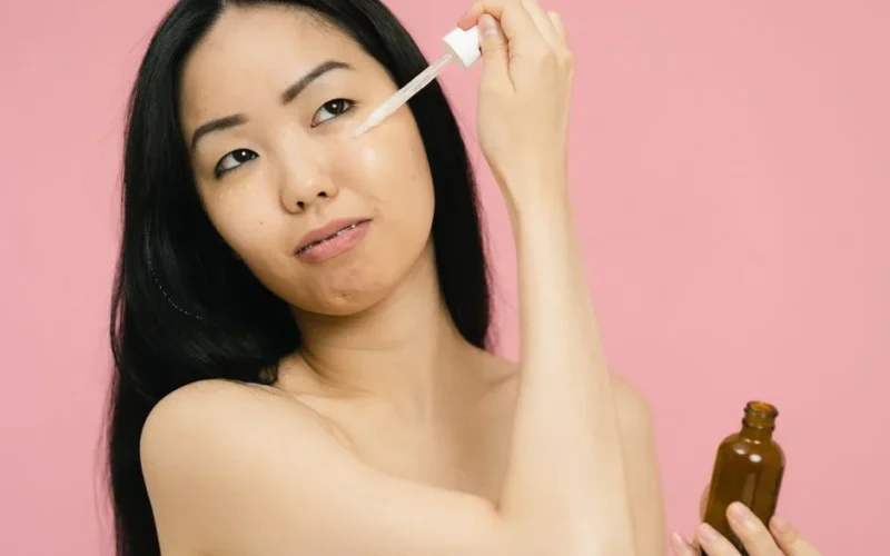 Asian woman applying skincare serum, holding a bottle against a pink backdrop indoors.