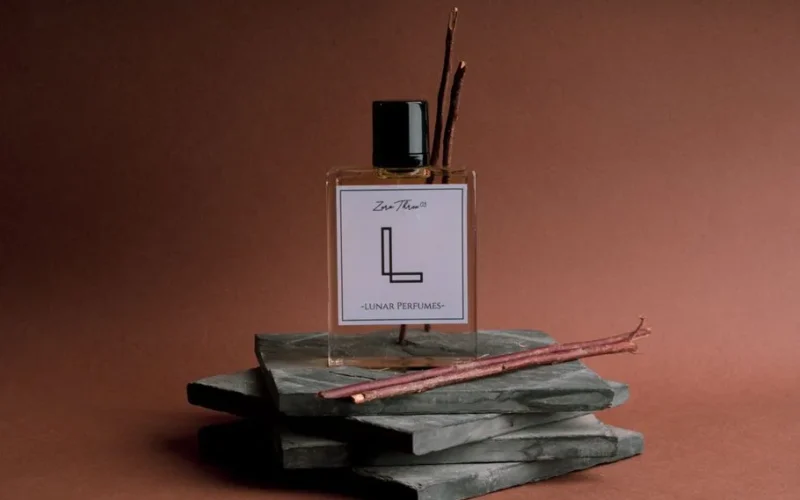 A still life featuring a perfume bottle and sticks on a slate platform against a brown background.