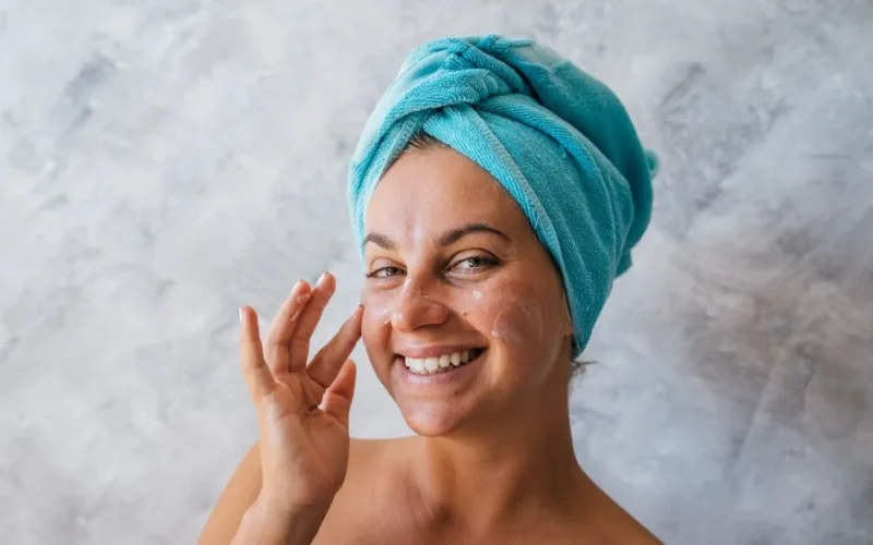 Happy woman with towel on head applies moisturizer, showcasing skincare routine.