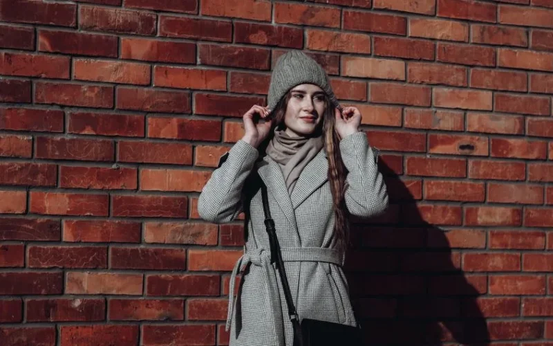 Stylish woman in gray coat and beanie standing against a brick wall.
