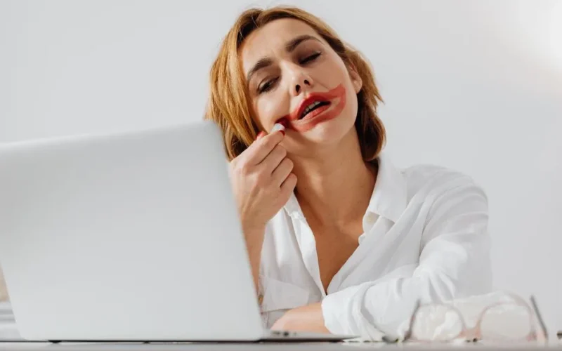 A woman with red lipstick smudged watching something on her laptop, indoors.