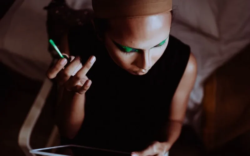 From above of crop ethnic female master applying eyeshadow on eyelid while looking at mirror