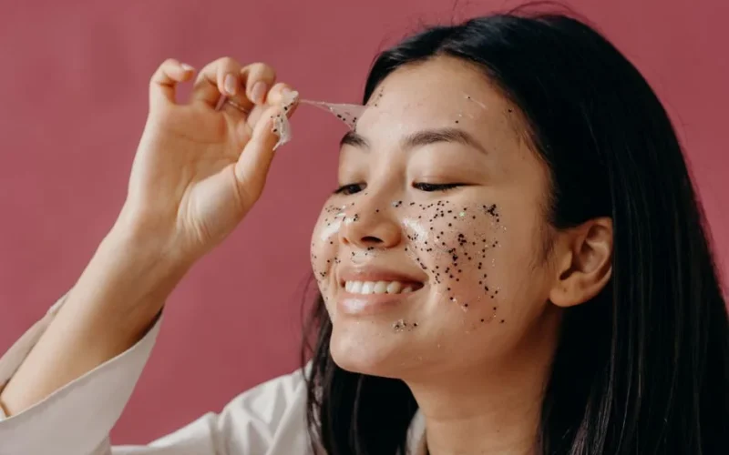 Close-up of smiling Asian woman peeling off a glittery face mask, highlighting her skincare routine.