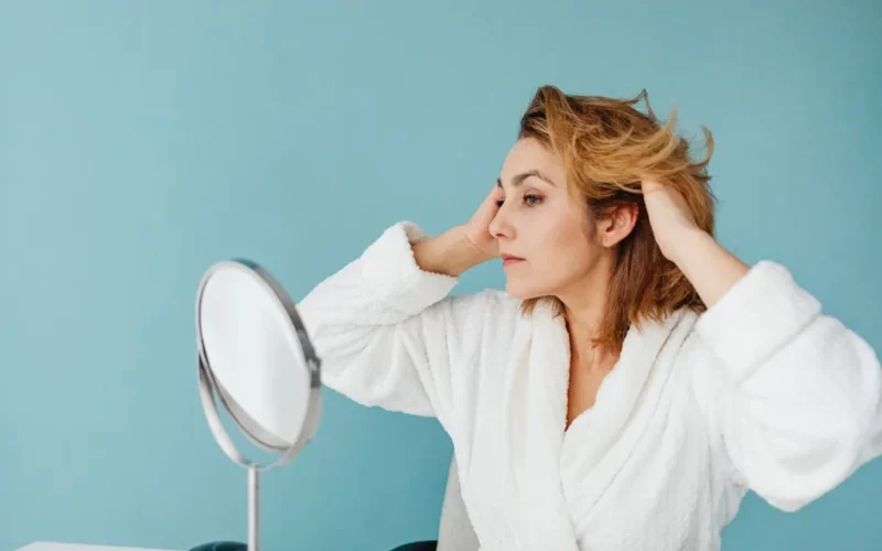 Adult woman in a bathrobe fixing her hair in front of a mirror against a blue background.